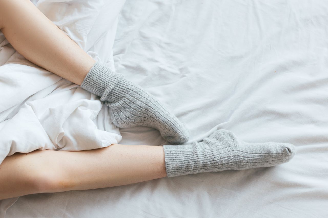 services-03 Adult feet wearing gray socks on a bed with white linens, evoking comfort and relaxation.