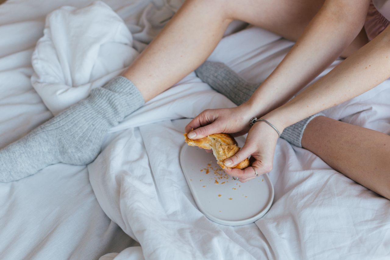 High angle of crop anonymous lady in warm socks sitting on soft bed and eating fresh yummy croissant in sunny morning at home